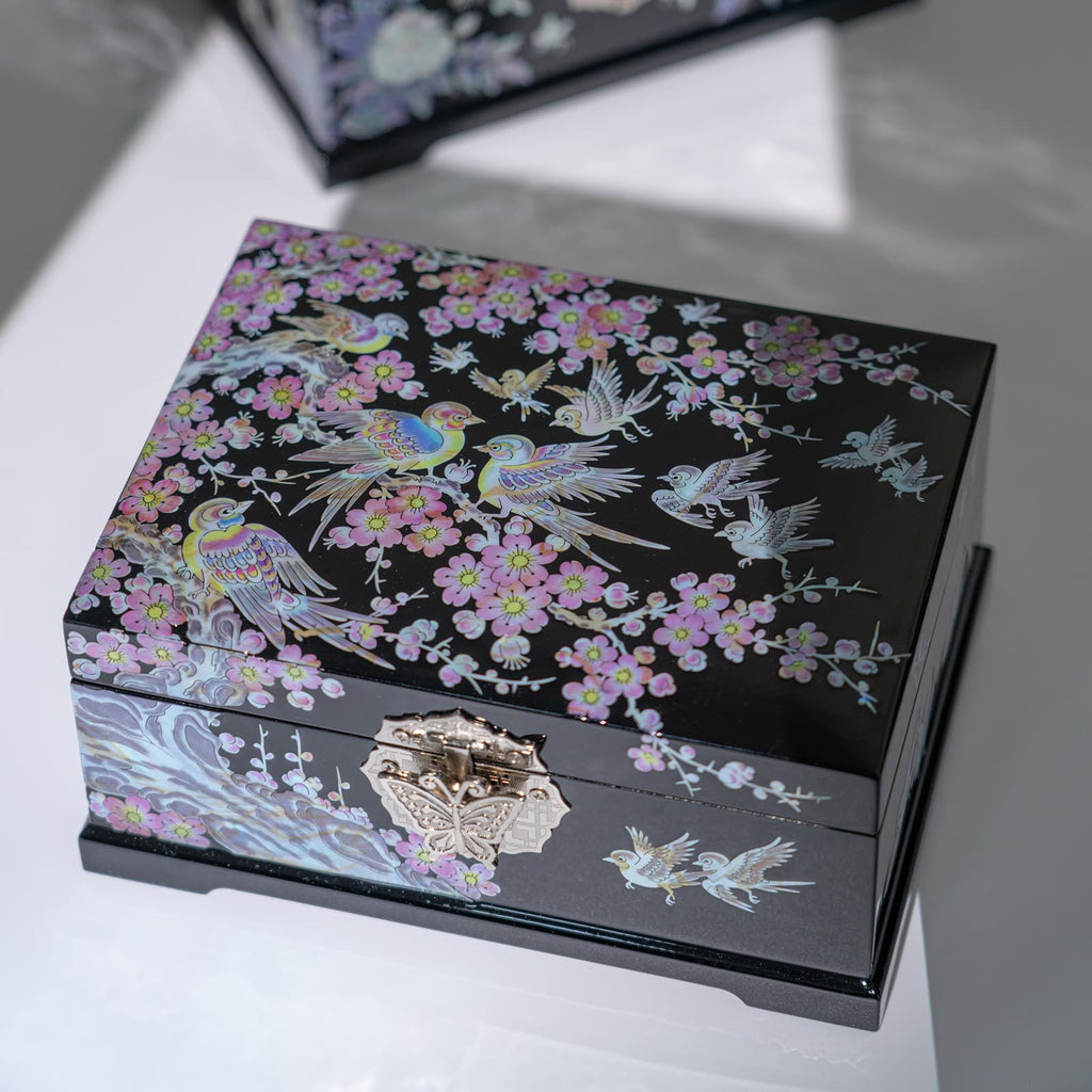 A black jewelry box with a mother of pearl inlay of birds and plum blossoms, featuring a butterfly clasp, placed on a reflective surface with a soft shadow.
