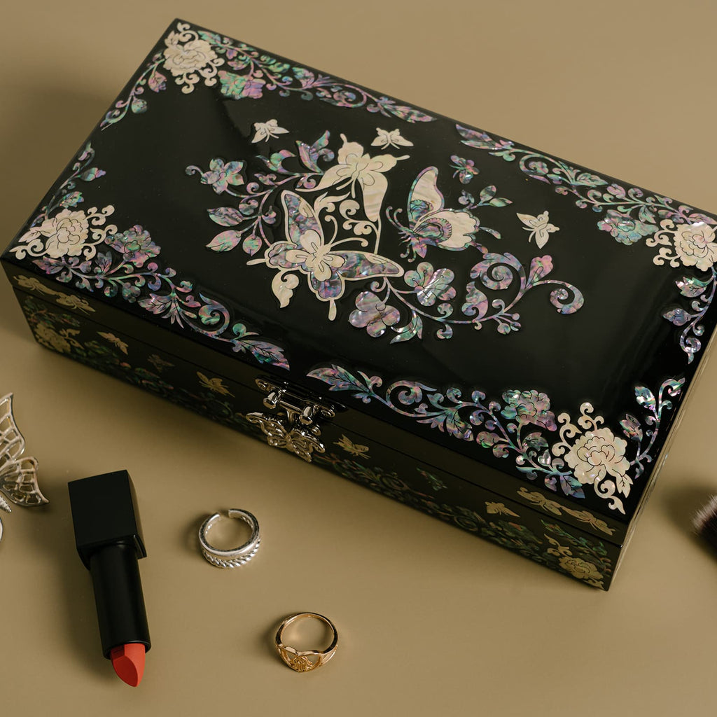 A black lacquered jewelry box with a radiant mother-of-pearl butterfly and floral design is presented against a neutral backdrop, with a tube of red lipstick, two rings, and a silver earring adding a touch of elegance to the composition.