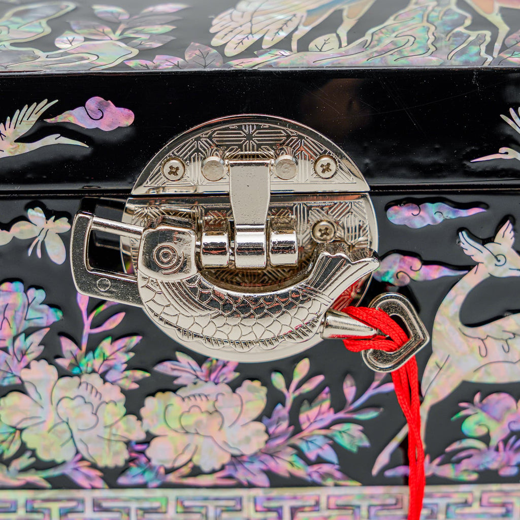 A close-up of a black lacquered jewelry box latch with a red cord, showcasing intricate mother-of-pearl inlay work with delicate floral and bird motifs in the background.