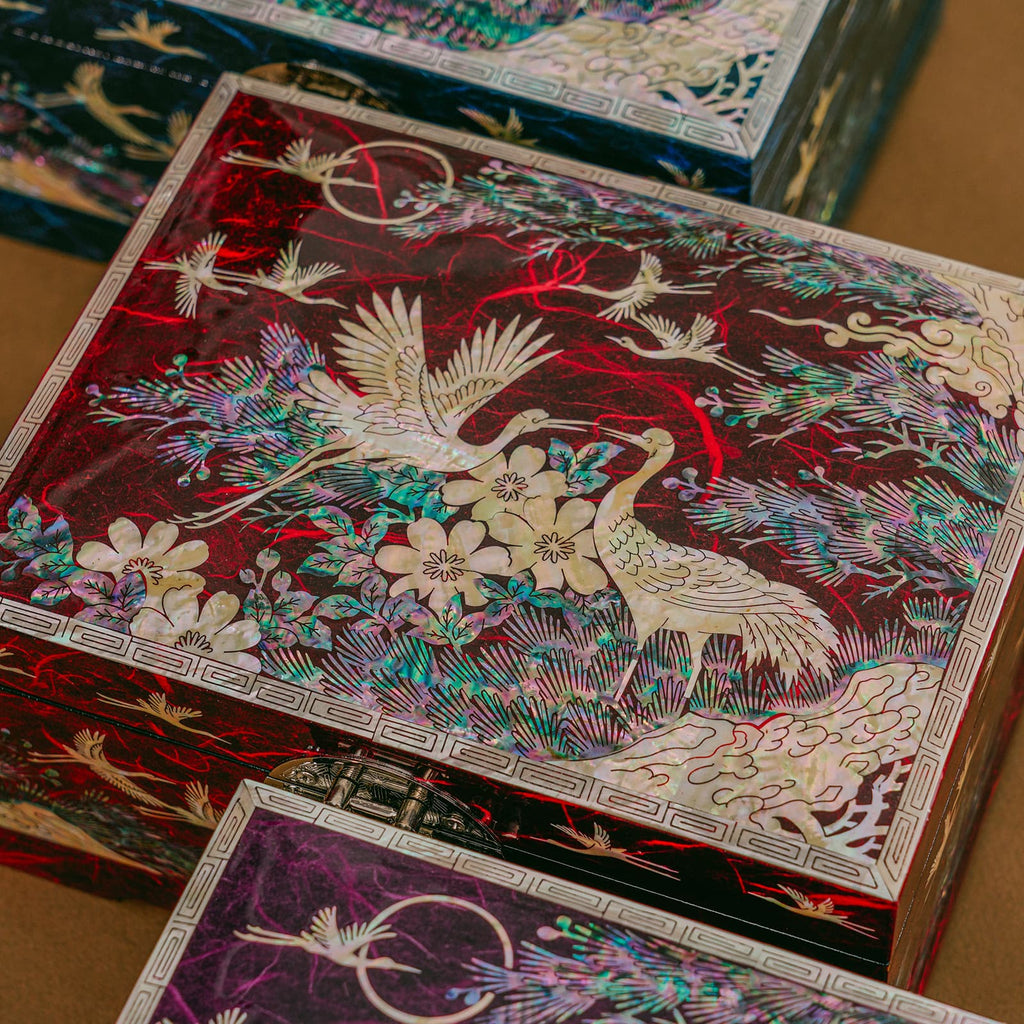 A close-up of a collection of Mother of Pearl boxes, the nearest one showcasing a vibrant red background with cranes and floral designs.