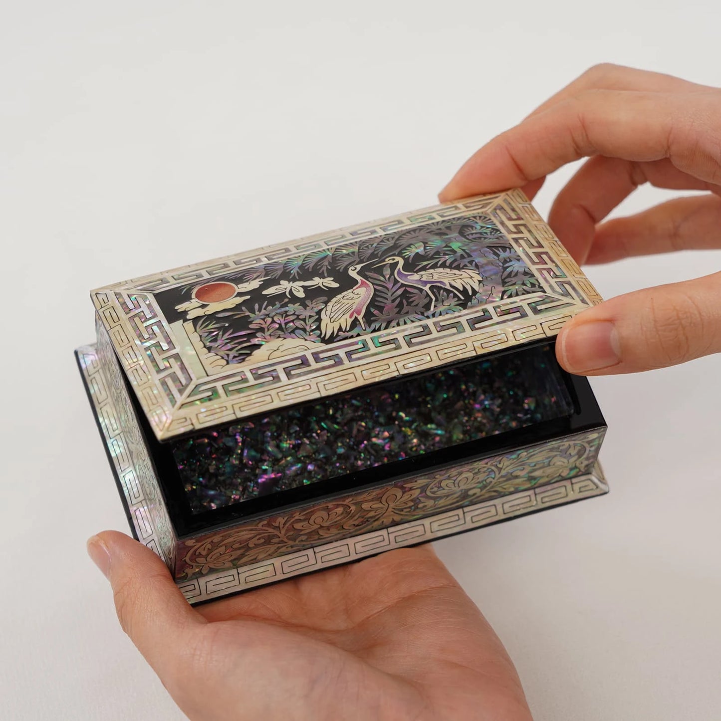 A close-up of a handcrafted mother of pearl lacquer box being opened, showing a detailed crane and pine tree scene (Sipjangsaeng