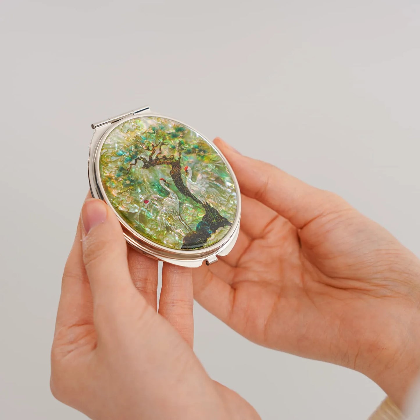 A compact mirror featuring white cranes and a tree with green foliage, set against a shimmering background.