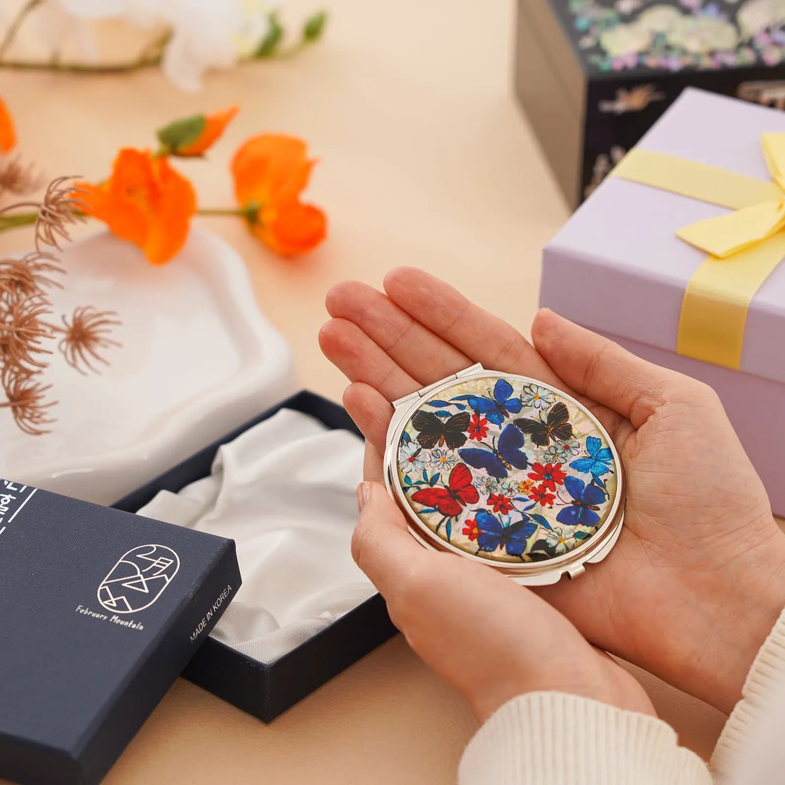 A compact mirror with a colorful butterfly and floral design, placed inside an open navy blue gift box