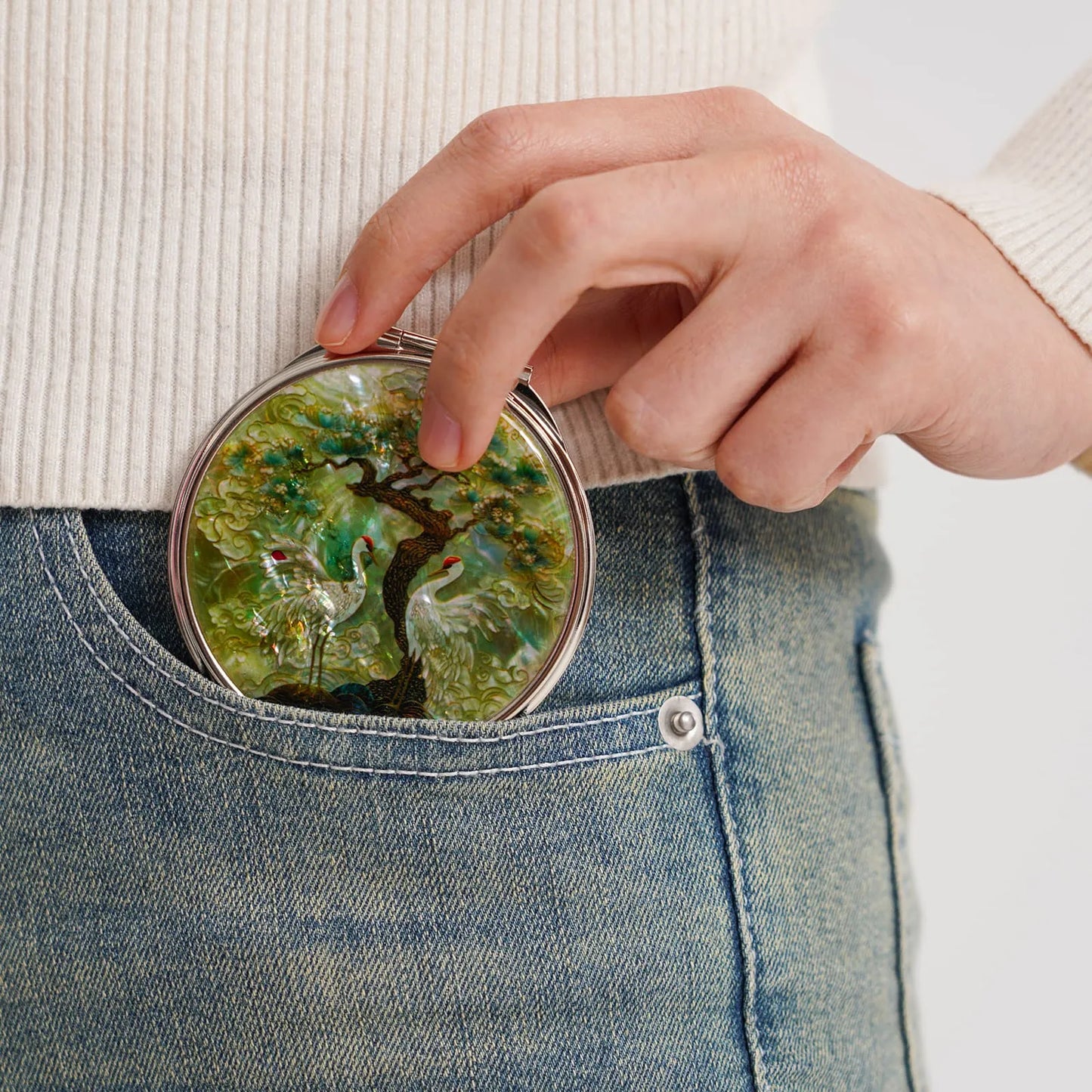 A compact mirror with a crane and tree design, partially tucked into a blue jean pocket