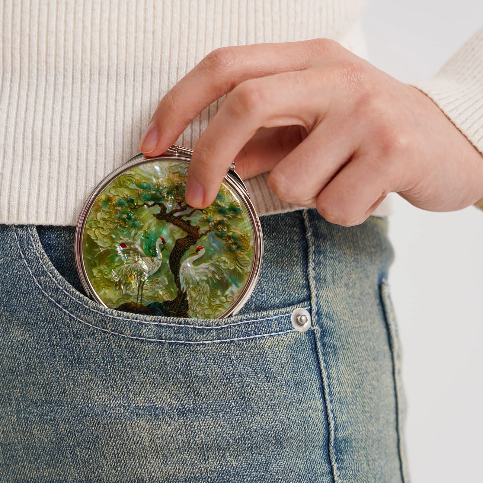 A compact mirror with a crane and tree design, partially tucked into a blue jean pocket