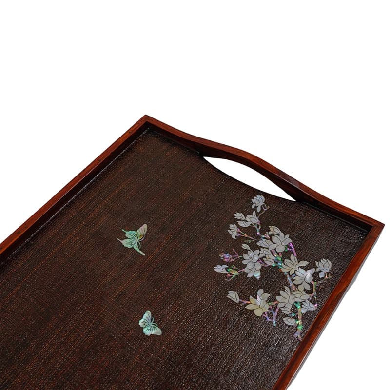 A corner view of a wooden tray with mother-of-pearl inlaid florals and butterfly, with handle.