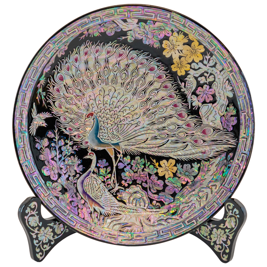 A decorative plate showcasing a peacock with an expanded tail amid flowers, displayed on a stand with mother-of-pearl details.
