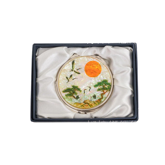 A decorative round item featuring a bright orange sun, birds in flight, and trees, placed inside a satin-lined box