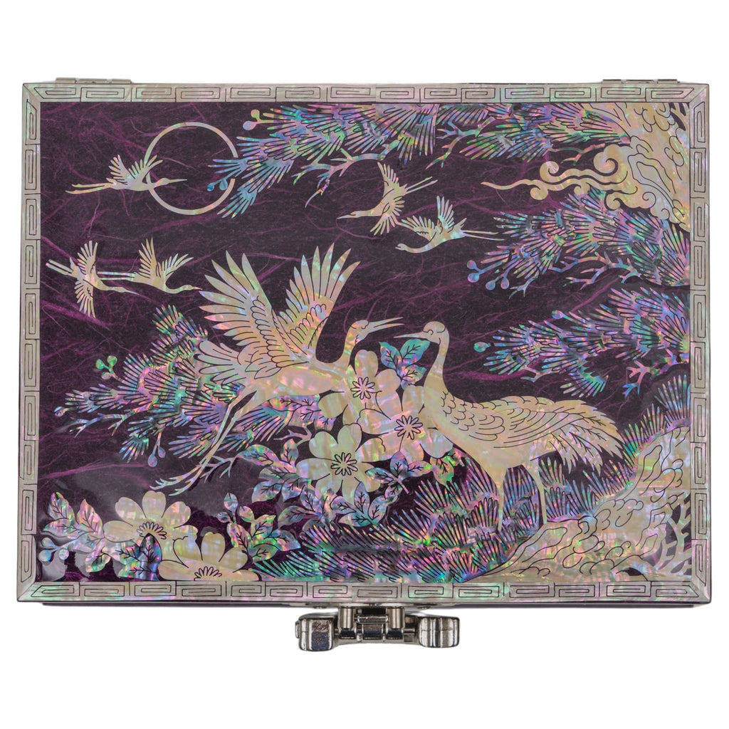 A flat-lay view of a closed mother-of-pearl inlaid box, featuring vibrant crane and floral motifs on a purple background with a metallic front latch.