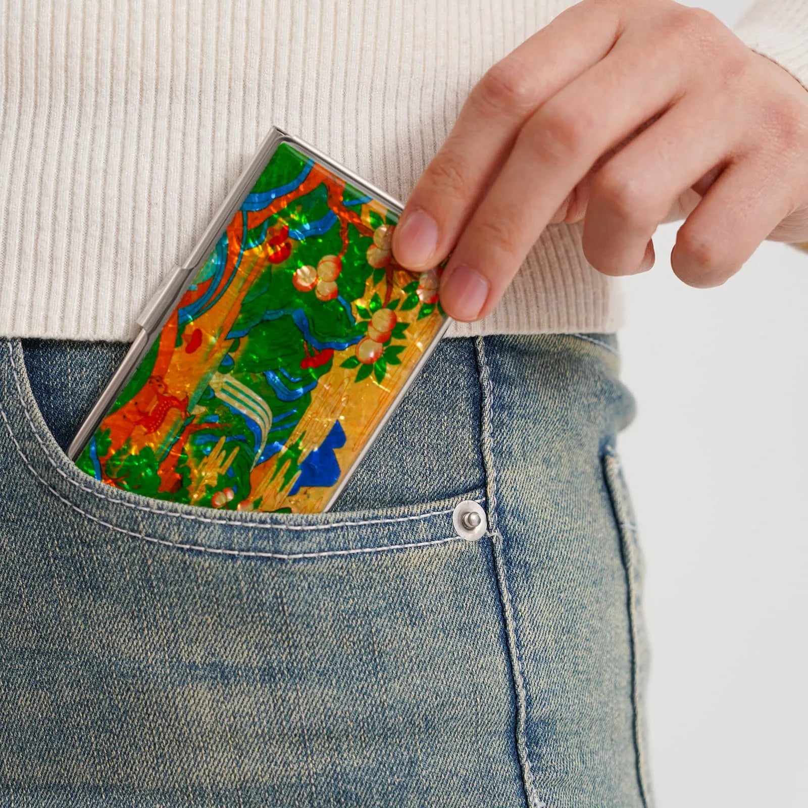A hand places a colorful case with floral and abstract patterns into the front pocket of blue jeans