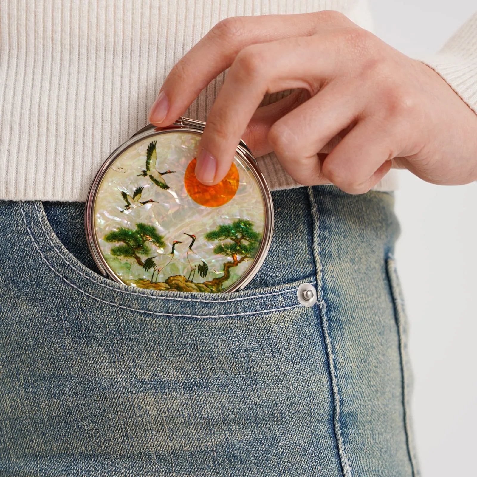 A hand placing a round compact mirror into the front pocket of blue jeans. The mirror features cranes, trees, and a bright orange sun in its design