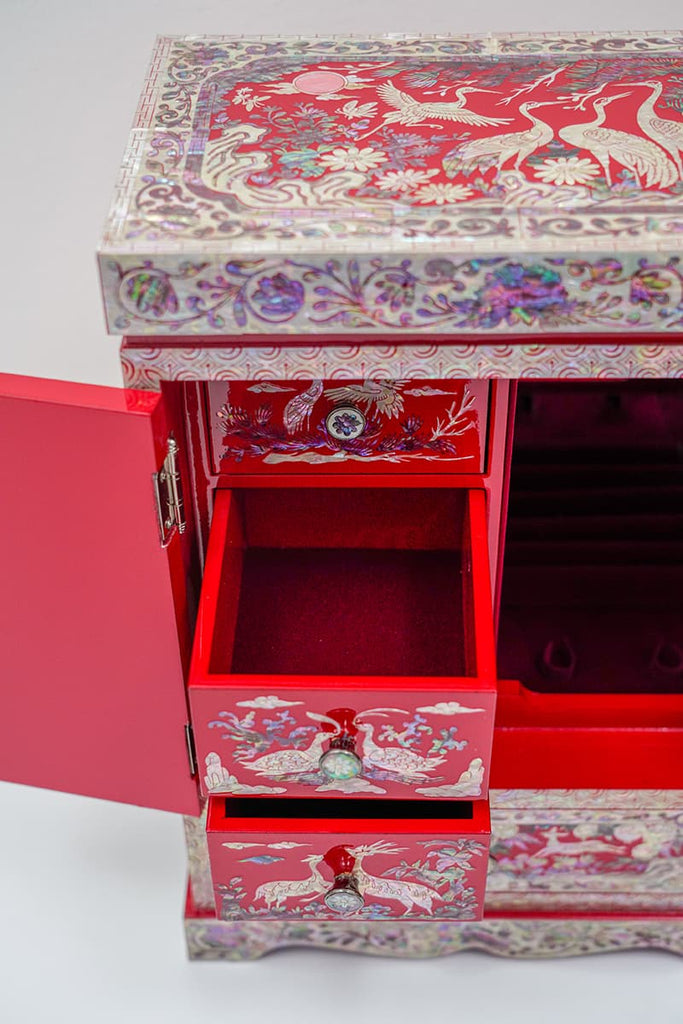 The top of a jewelry box with mother of pearl inlay, showcasing cranes and floral designs on a red backdrop, surrounded by a detailed border.A jewelry box with an open drawer, revealing a red velvet interior, and mother of pearl inlay featuring cranes on the exterior.