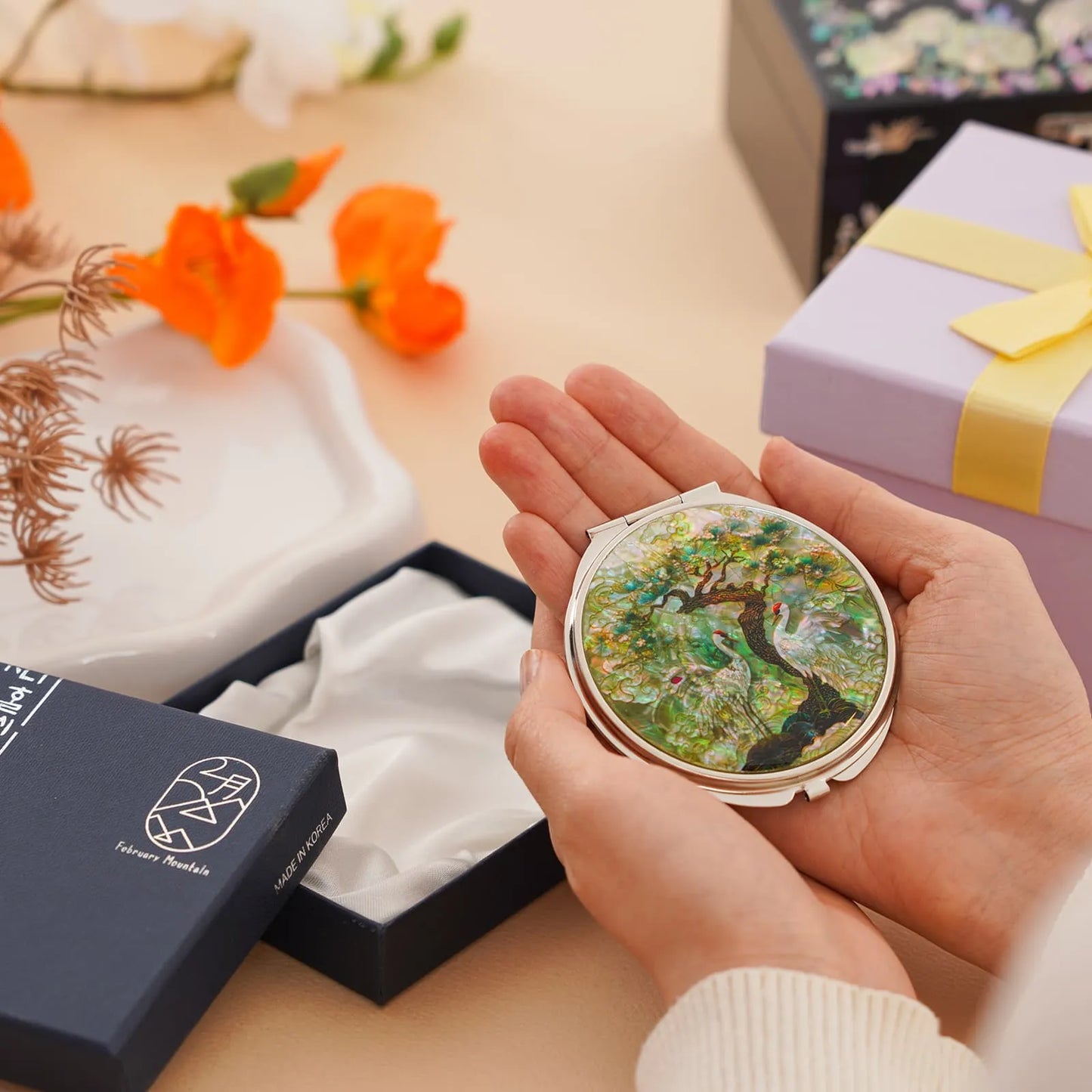 A person's hands holding a compact mirror featuring a tree and bird design, above an open satin-lined gift box