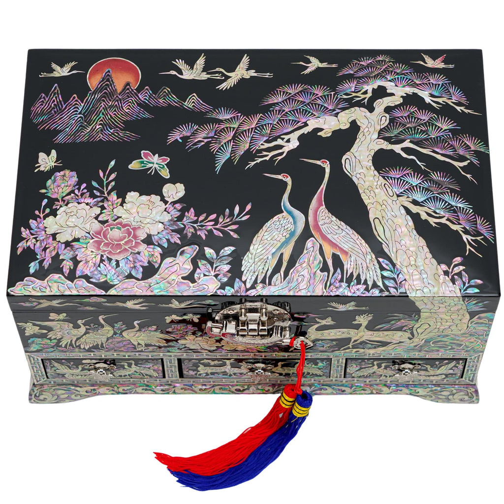 A rectangular mother-of-pearl box depicting a serene landscape with cranes and a rising sun, accented with a vibrant, traditional Korean norigae tassel hanging from the latch.