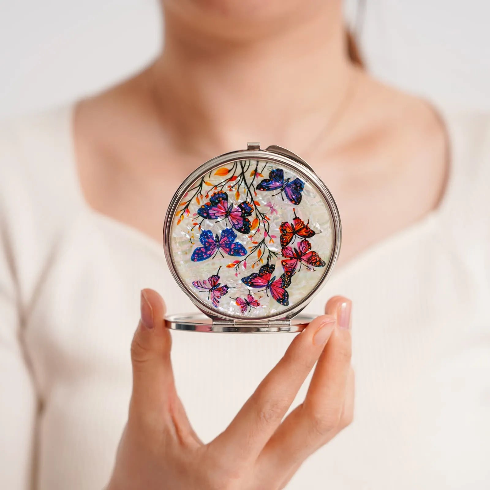 A round compact mirror featuring a delicate design of butterflies in shades of blue, pink, purple, and orange, resting on branches against a light backgroun