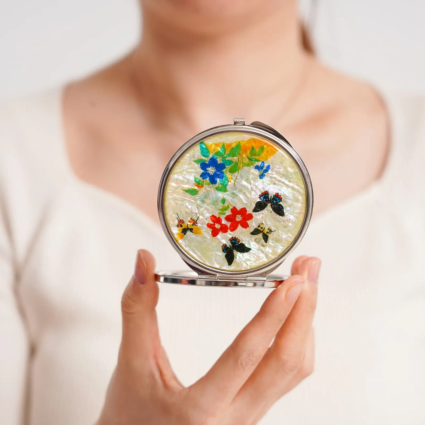 A round compact mirror featuring colorful flowers and leaves with an iridescent finish, held close to the camera