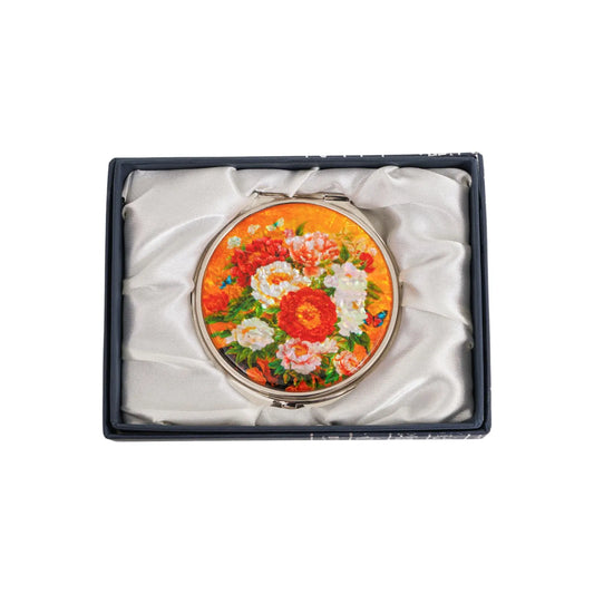 A round compact mirror with a colorful floral design featuring red, white, and orange flowers against a yellow background, placed inside a black satin-l