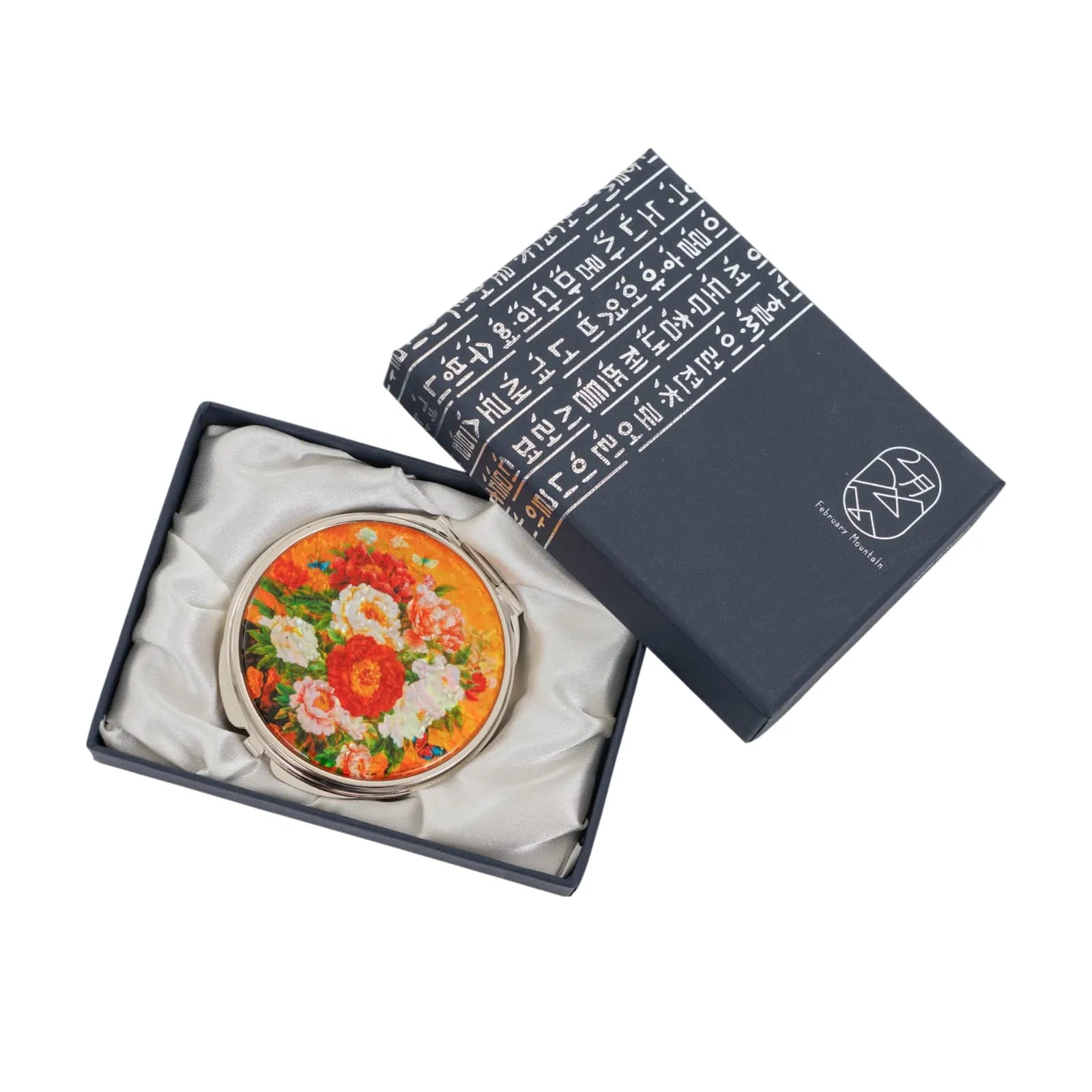A round compact mirror with a floral design featuring red, pink, and white flowers on an orange background, placed inside a satin-lined dark blue box wi