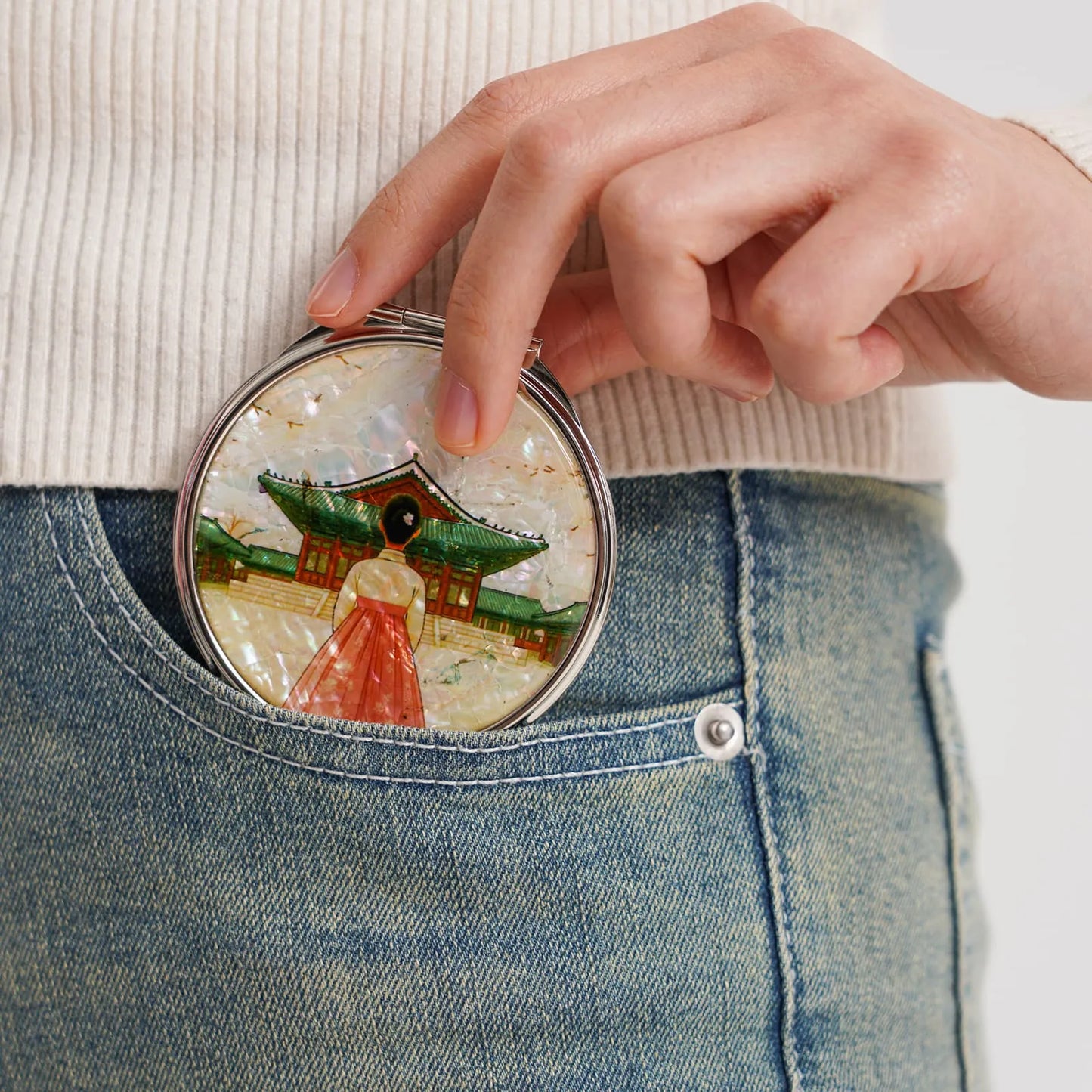 A round compact mirror with an intricate design of a traditional building and a figure in front, tucked into jeans
