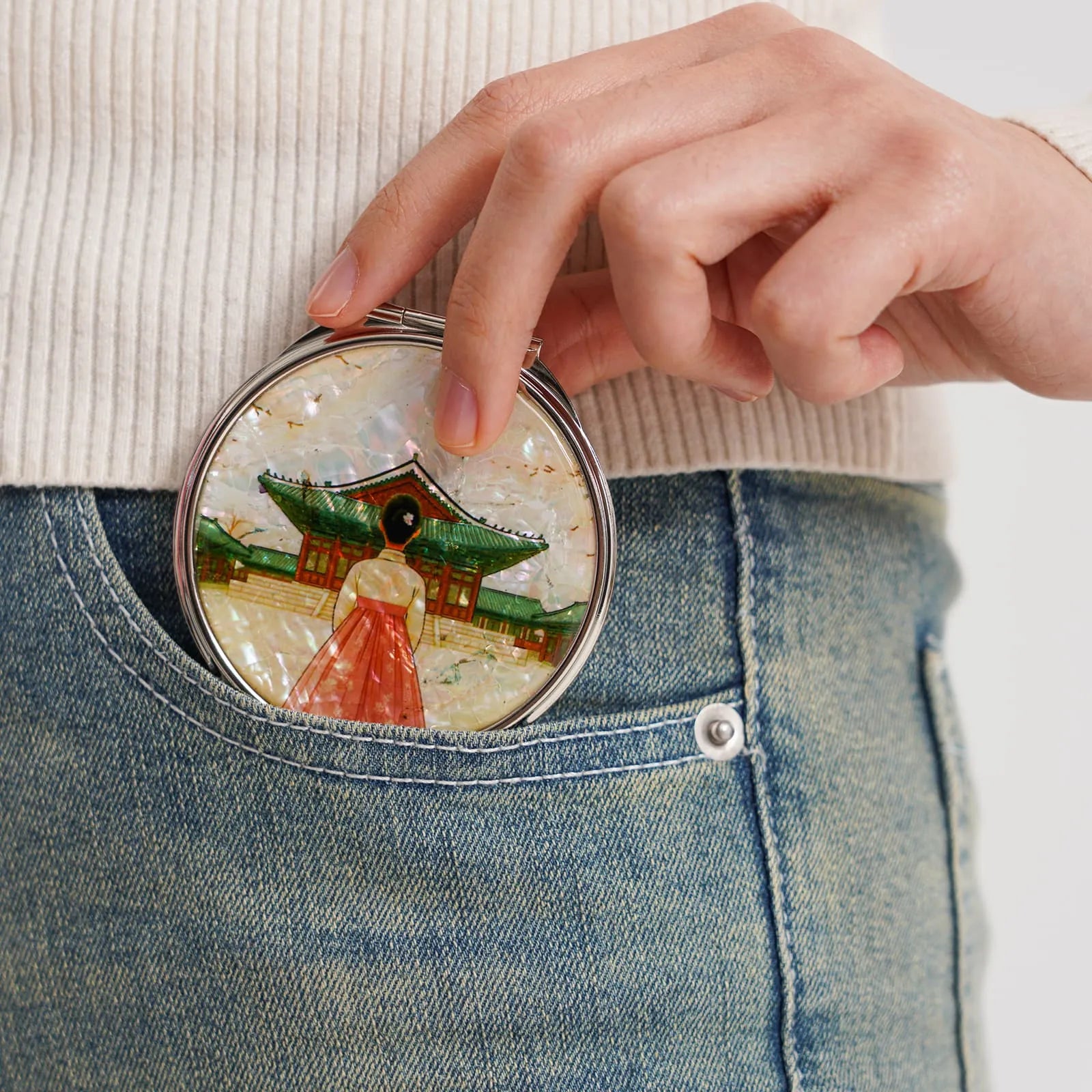 A round compact mirror with an intricate design of a traditional building and a figure in front, tucked into jeans