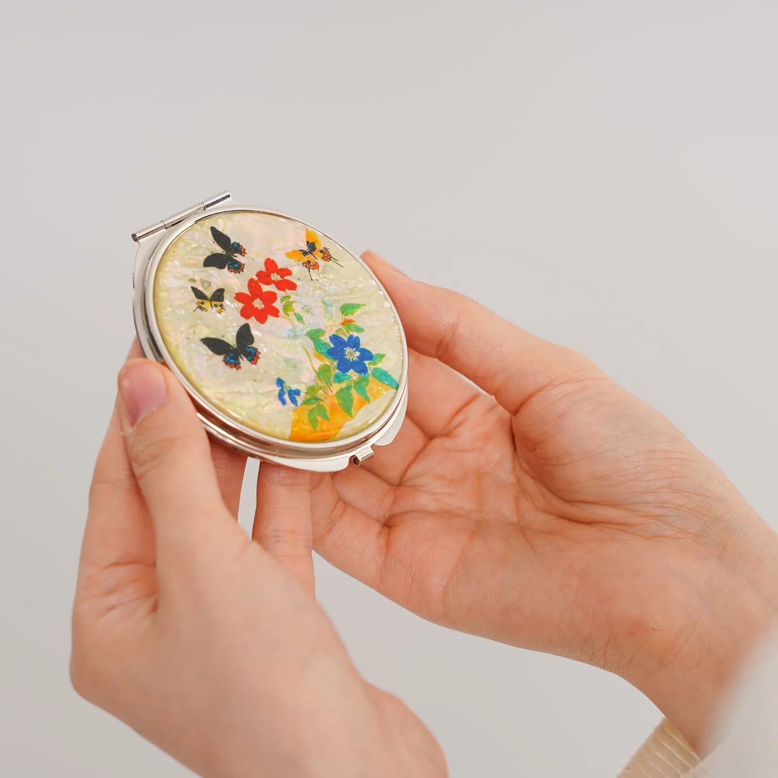 A round compact mirror with a pearlescent background, decorated with vibrant flowers and butterflies.