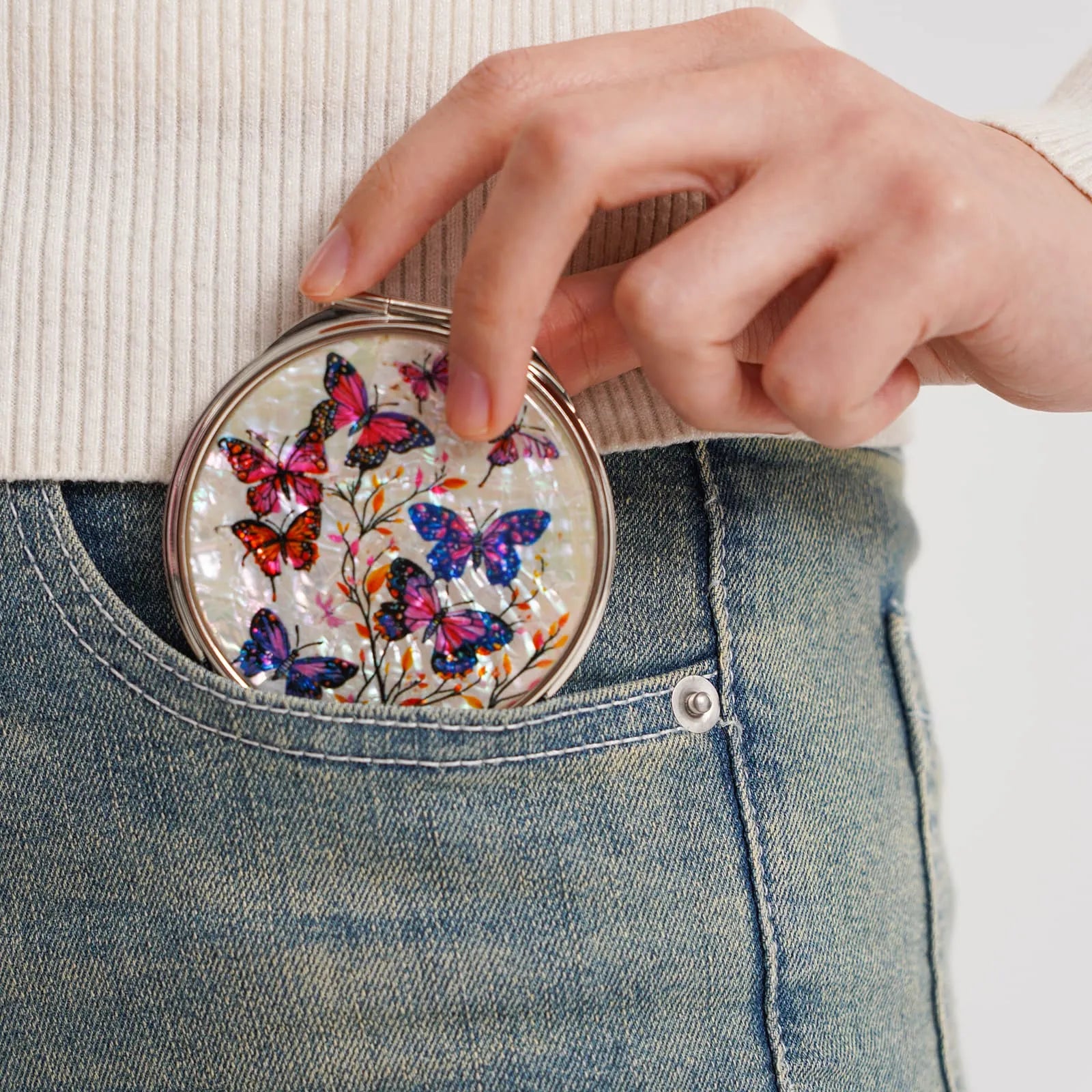 A round compact mirror with a vibrant butterfly design in shades of red, pink, purple, and blue, being placed into the front pocket of blue jeans