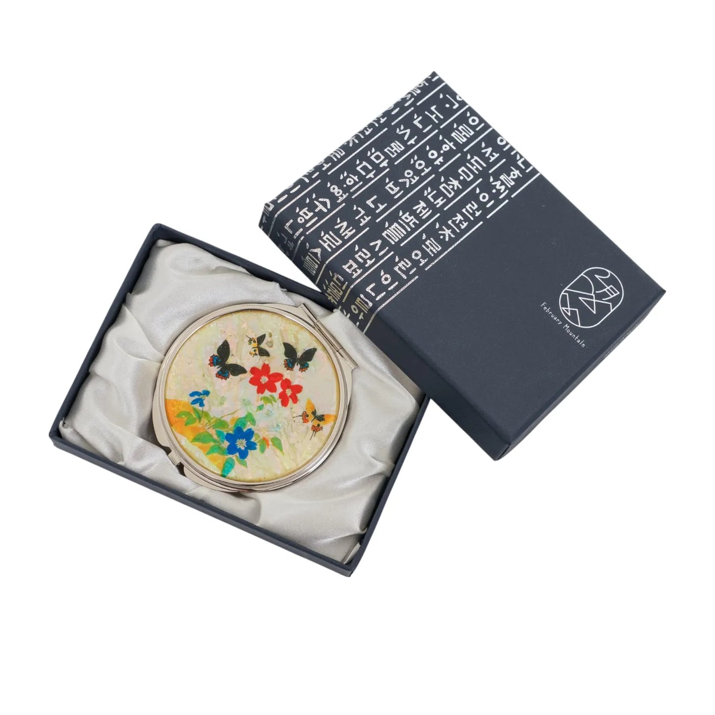 A round compact mirror with a vibrant floral and butterfly design, nestled in a dark blue gift box with Korean text