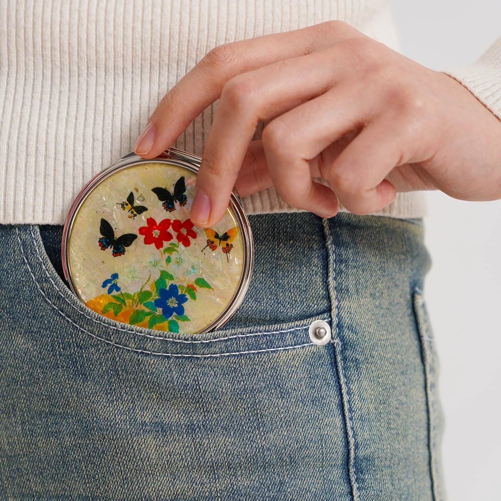 A round compact mirror with colorful butterfly and floral patterns, partially tucked into a blue jean pocket