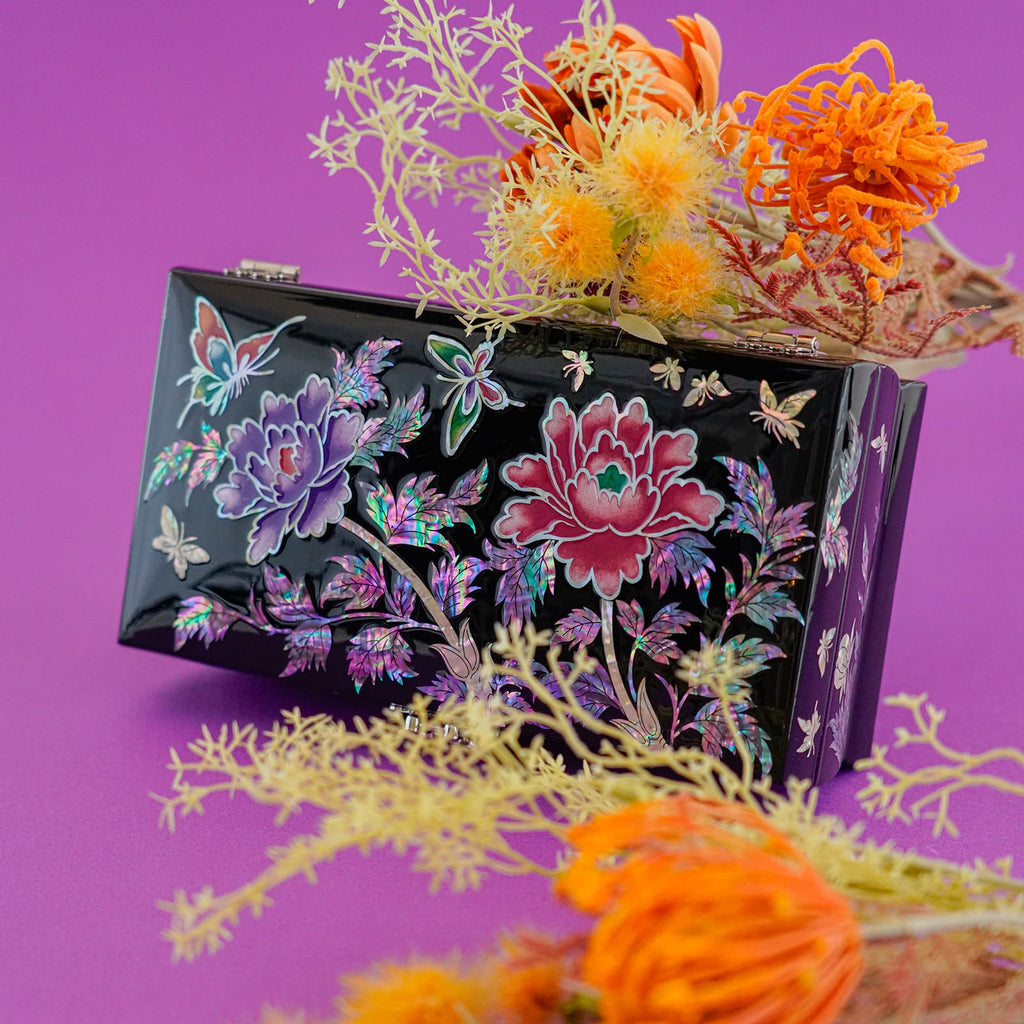 A small, intricately decorated mother-of-pearl clutch is showcased against a vibrant purple backdrop, complemented by colorful blooms that enhance its beauty.