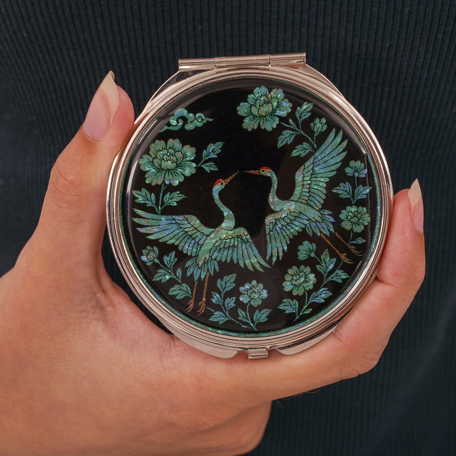 Black compact mirror with twin cranes and peonies in hand over dark background