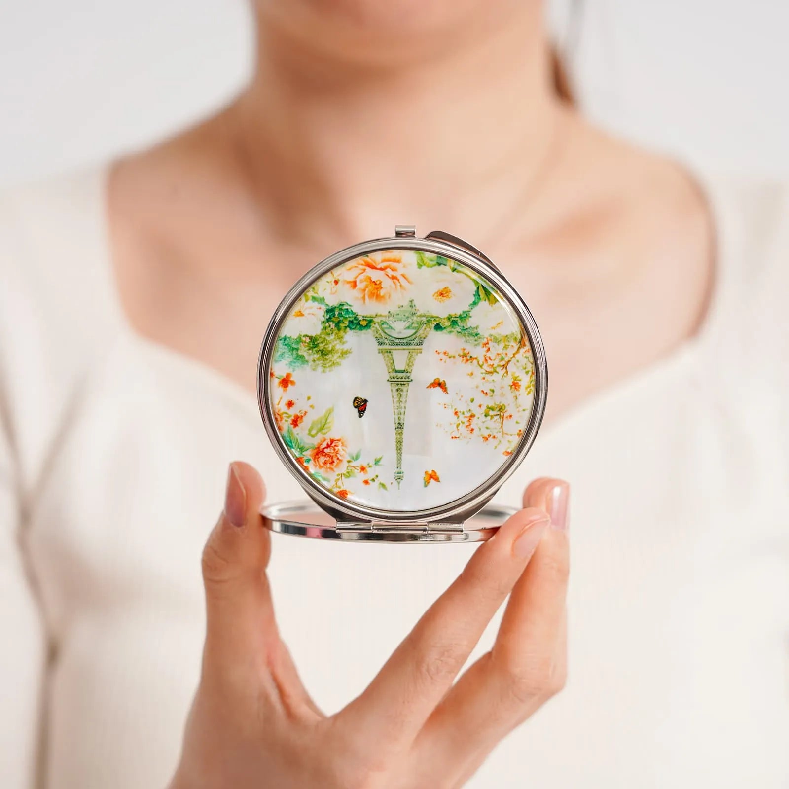 Compact mirror with elegant green and orange floral design, held by a person wearing a white top