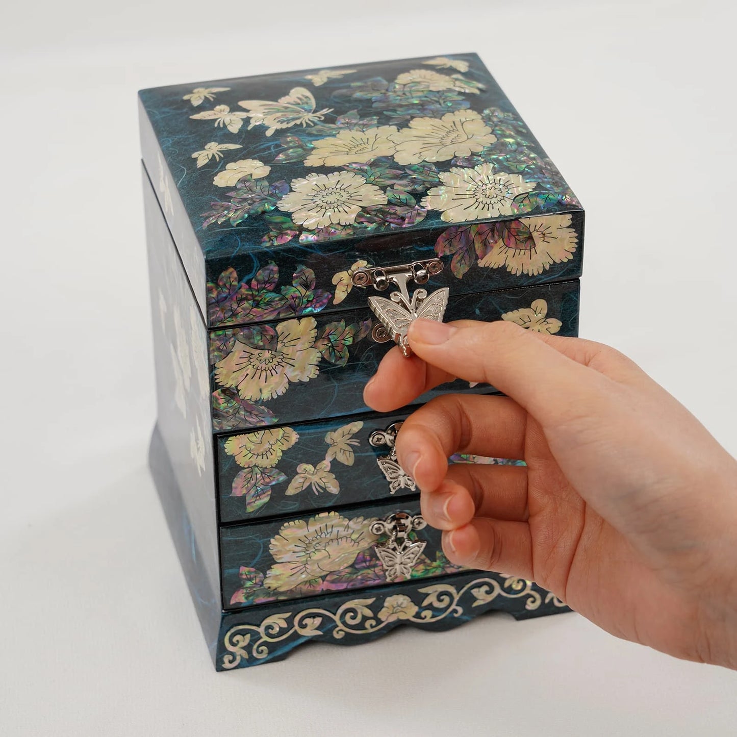 Detailed view of a hand opening a teal lacquer jewelry box. Showcasing iridescent butterfly locks and radiant artisan floral shell work.