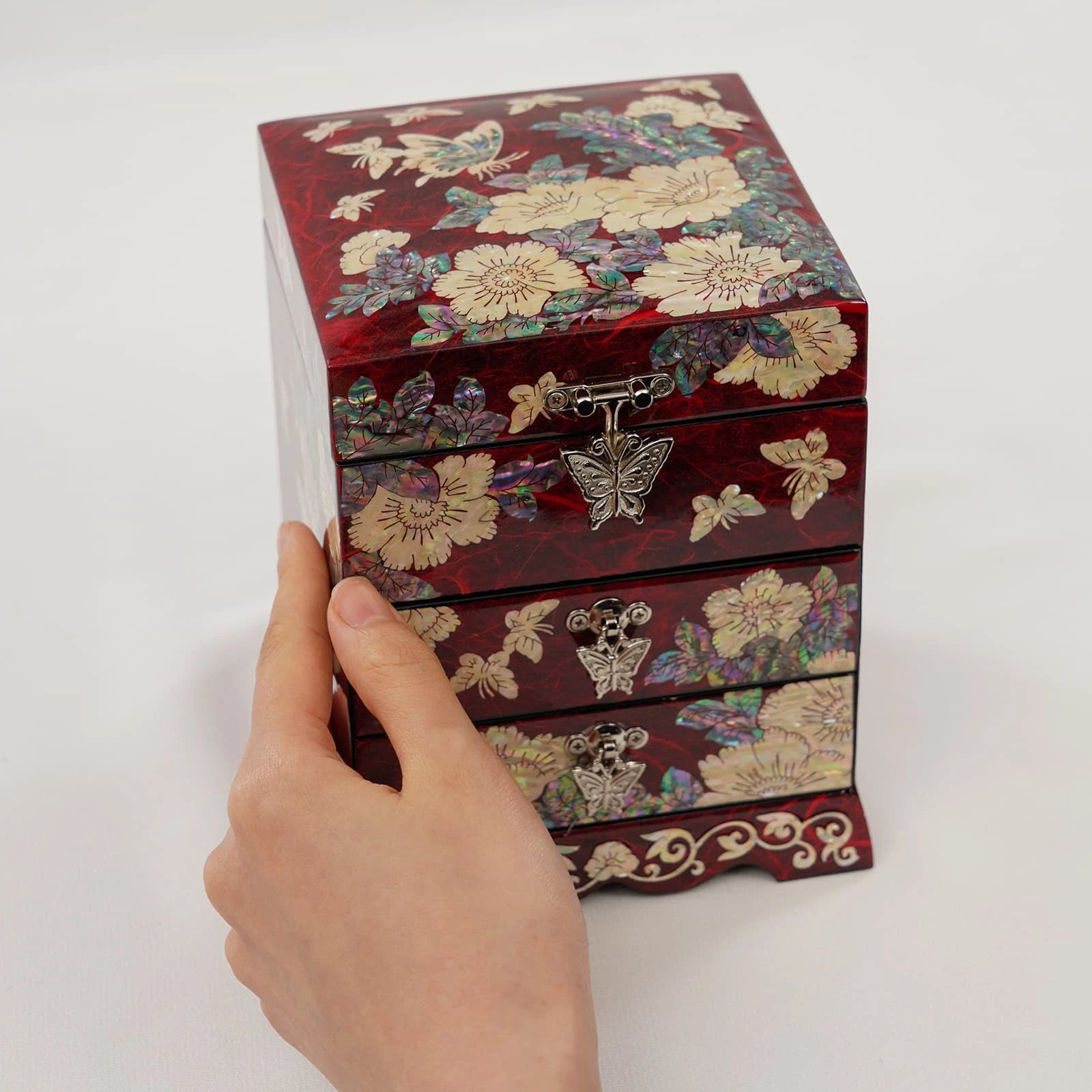 Hand holding a crimson red jewelry box with iridescent butterfly and peony inlay. Glossy lacquer finish storage for rings and vanity decor.