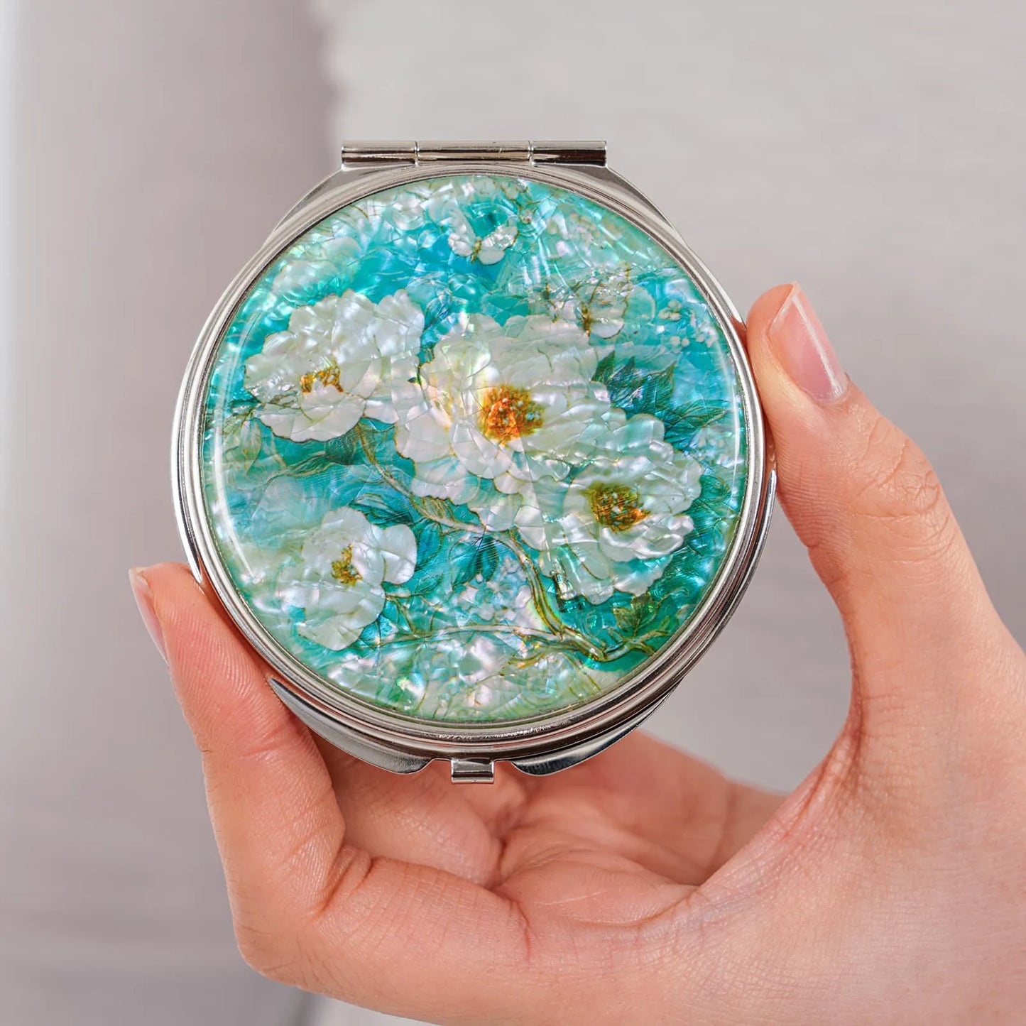 Hand holding round compact mirror with white peony and turquoise background mother-of-pearl
