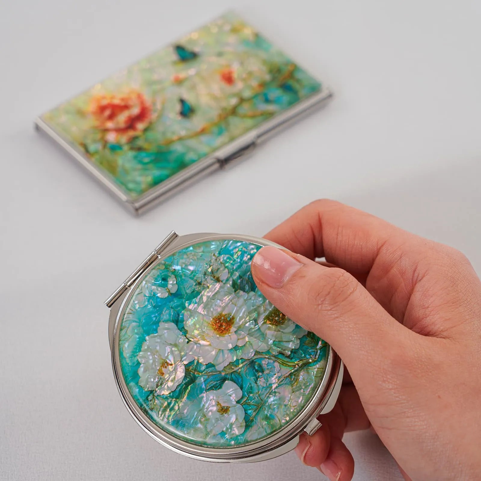 Hand holding round mother-of-pearl mirror with white flowers; floral card case in background