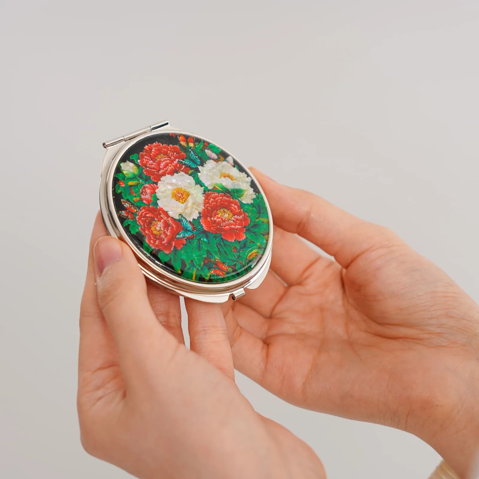 Hands holding a compact mirror with mother-of-pearl floral inlay on a plain background