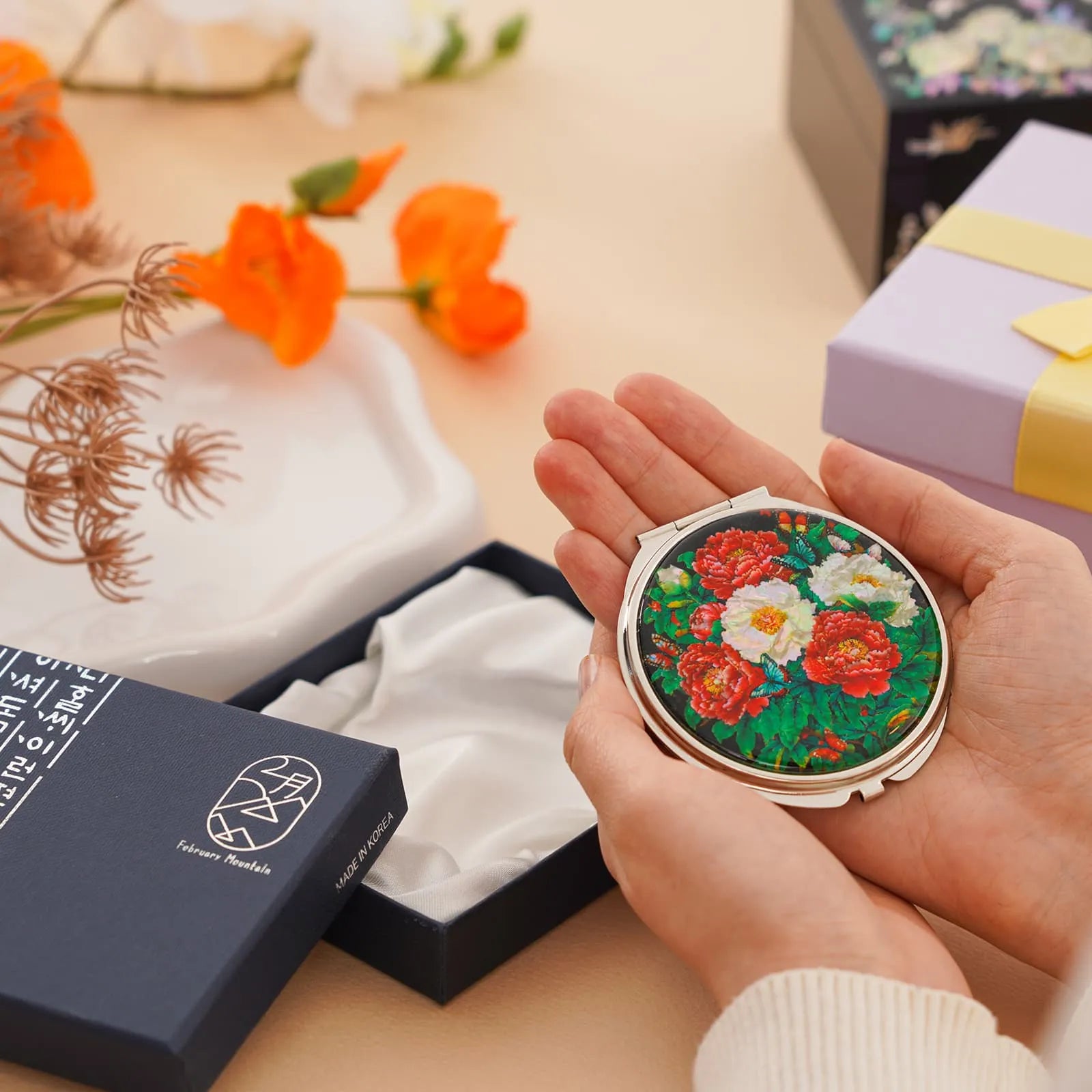 Hands holding floral compact mirror next to open gift box and orange flowers