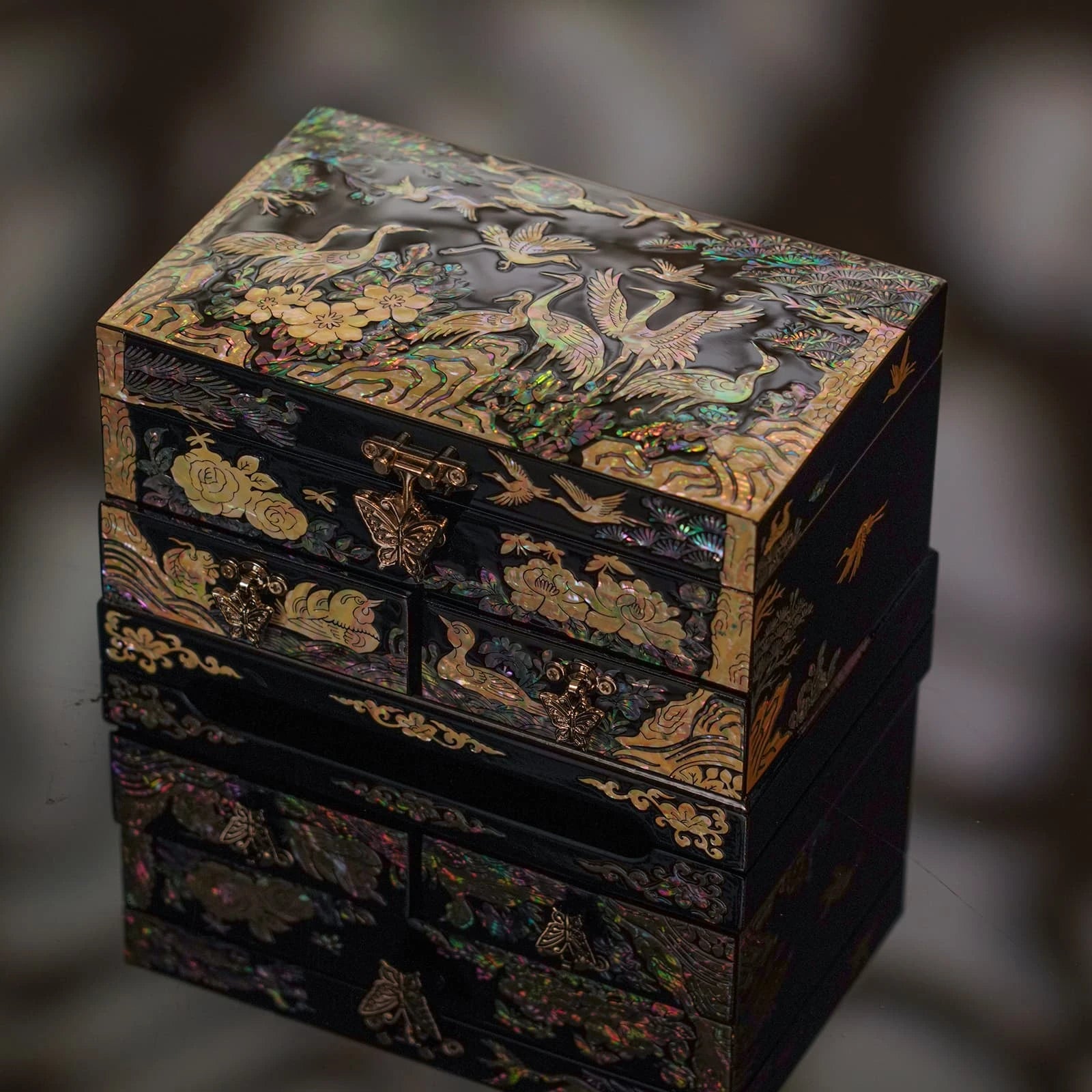 High-angle, reflective shot of a closed black nacre jewelry box. Intricate mother of pearl cranes and flowers shimmer against the dark surface and its reflection