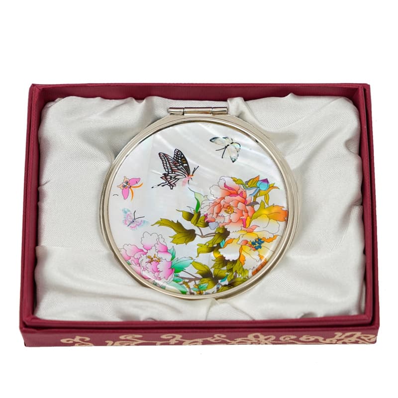  A round compact mirror with a floral and butterfly design, encased in a red box with a satin lining and decorative edging.