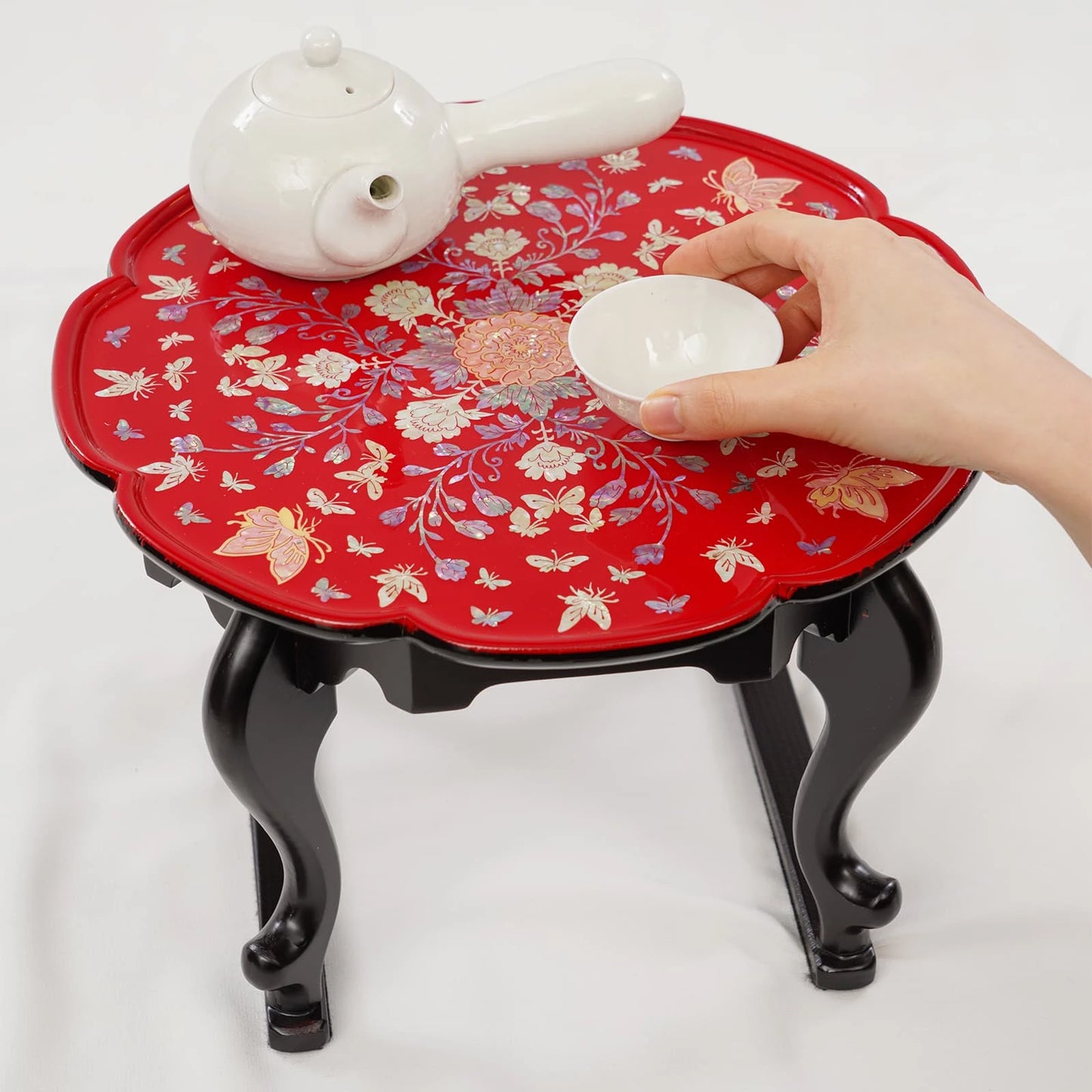 Red floral-patterned tray with black legs holding a teapot and a hand placing a white lid on it.