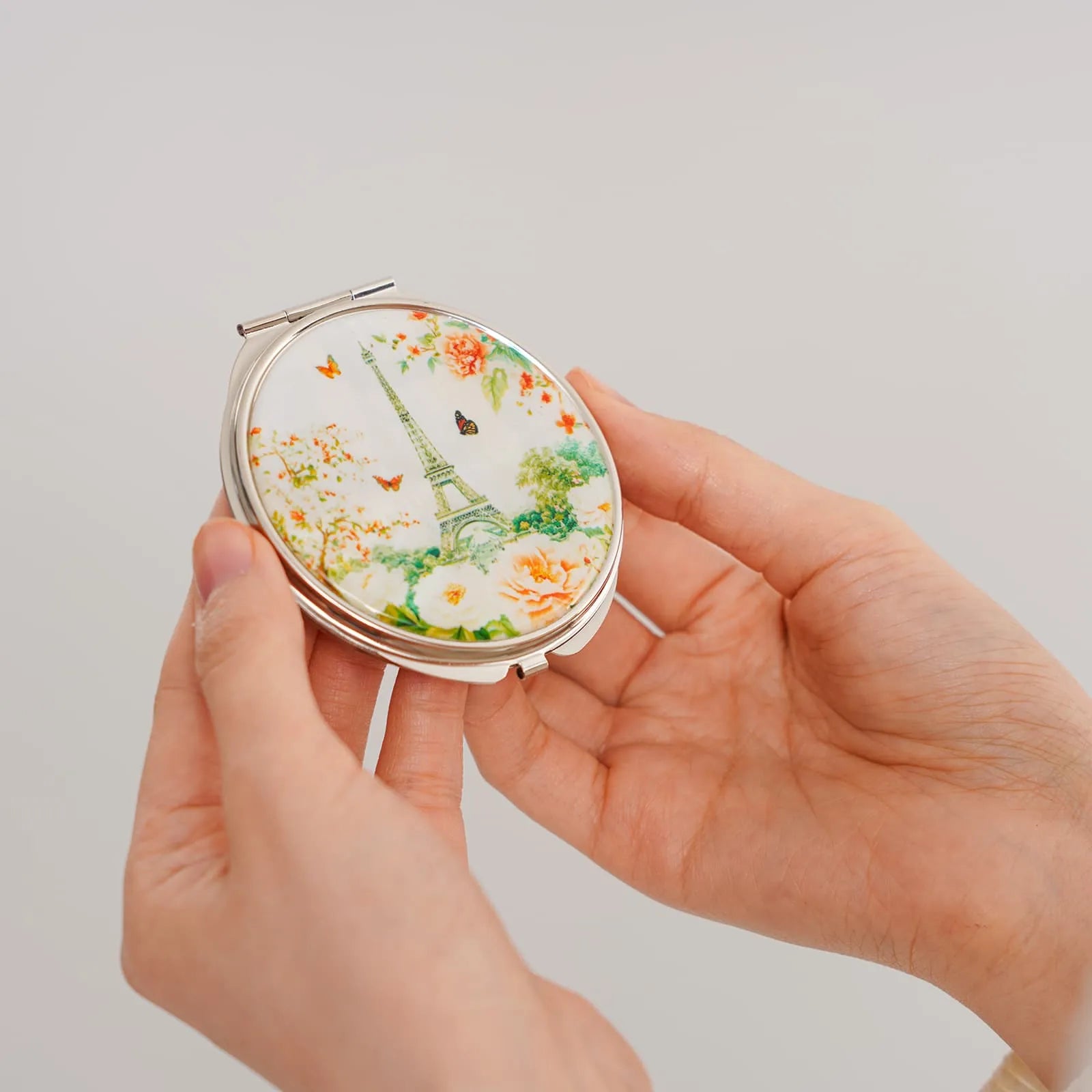 Round compact mirror with Eiffel Tower, flowers, and butterflies on the lid, held by hands against a light background