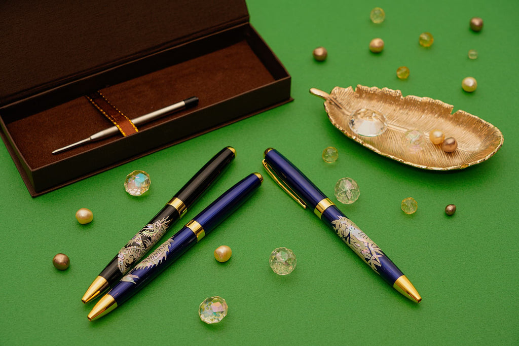 Three mother-of-pearl inlaid pens, a refill, and gold leaf dish rest on a green background.