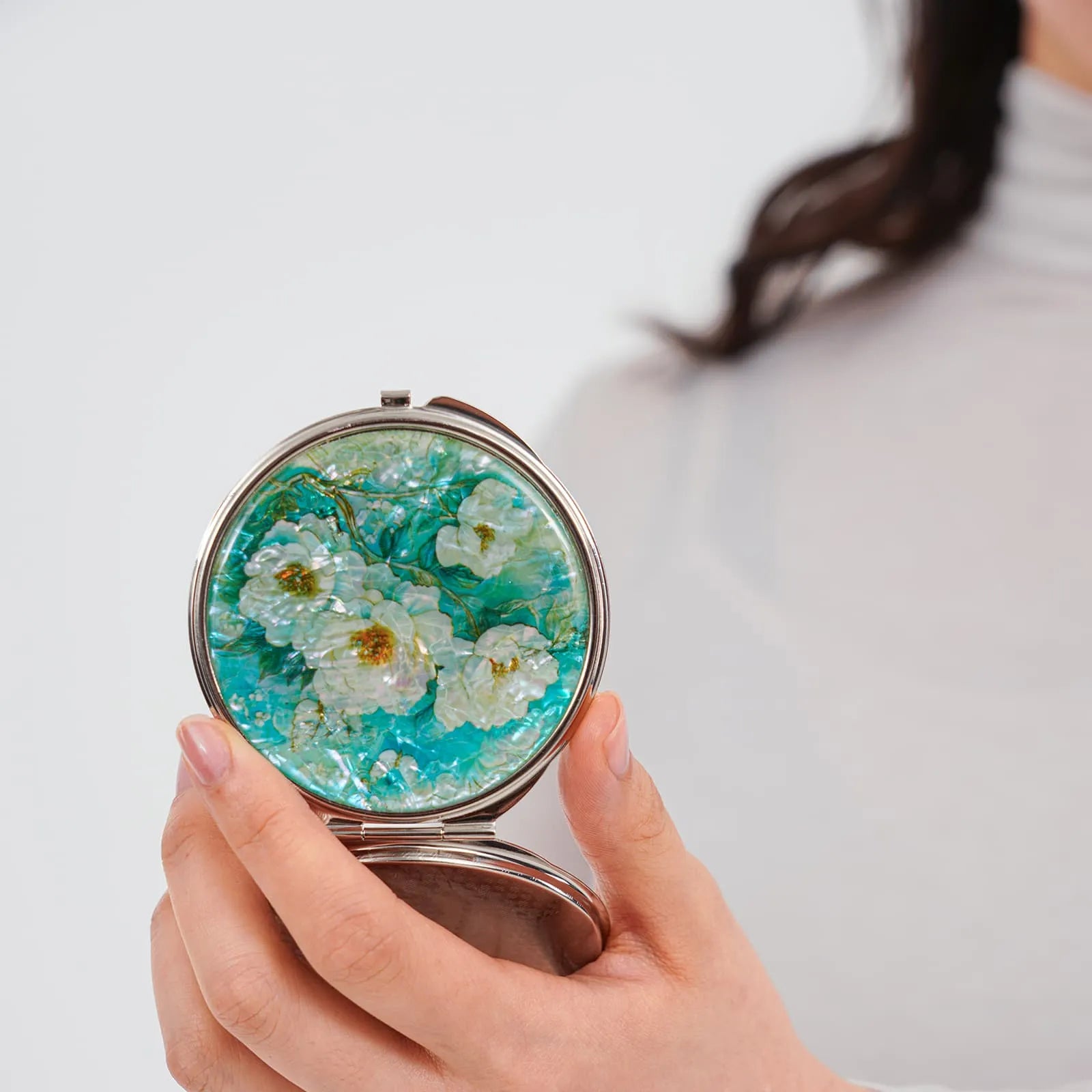Woman holding round compact mirror with floral and butterfly inlay in a turquoise background.