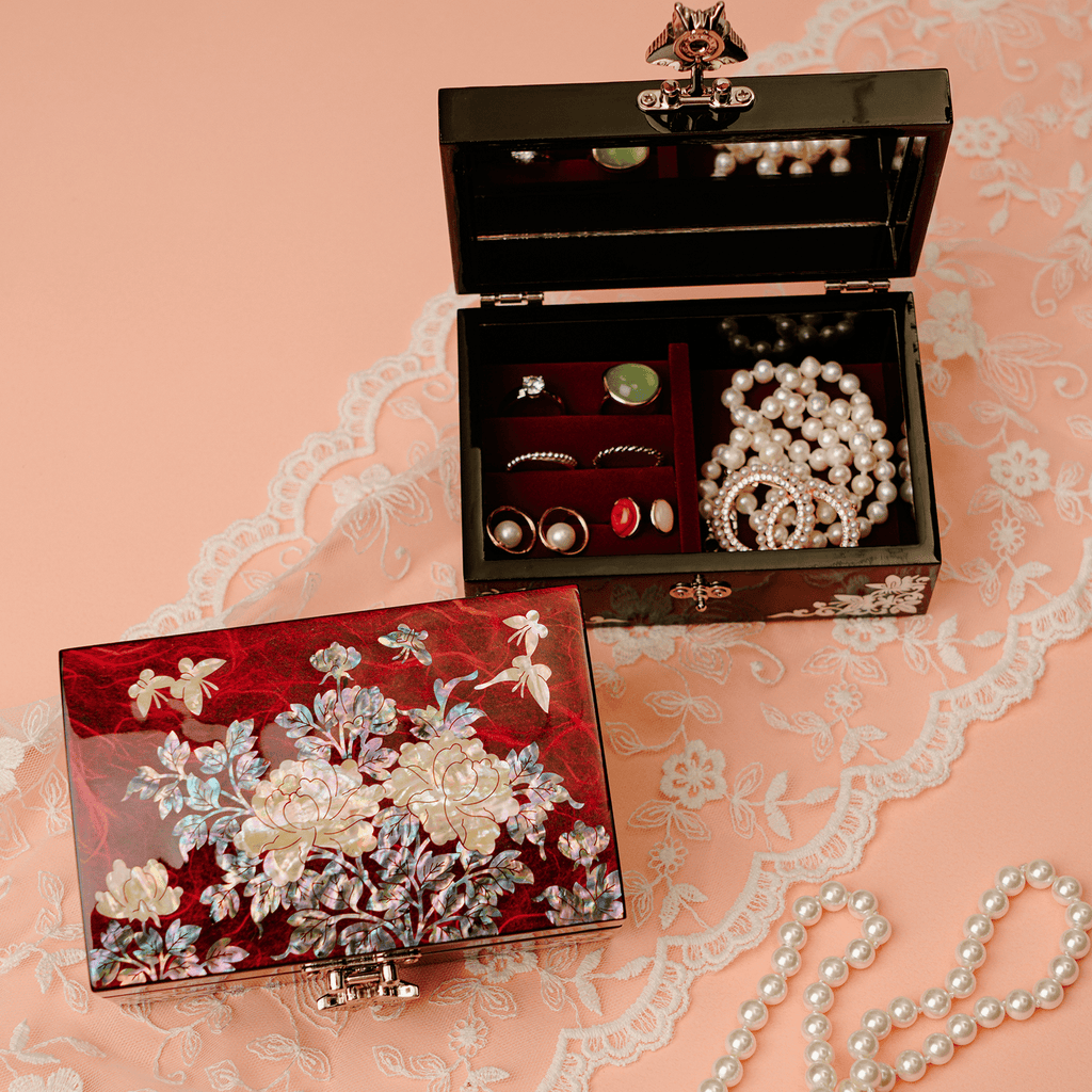  A red jewelry box with floral mother-of-pearl inlay, opened to reveal a black interior with a selection of jewelry on a red background, set against a delicate lace surface.