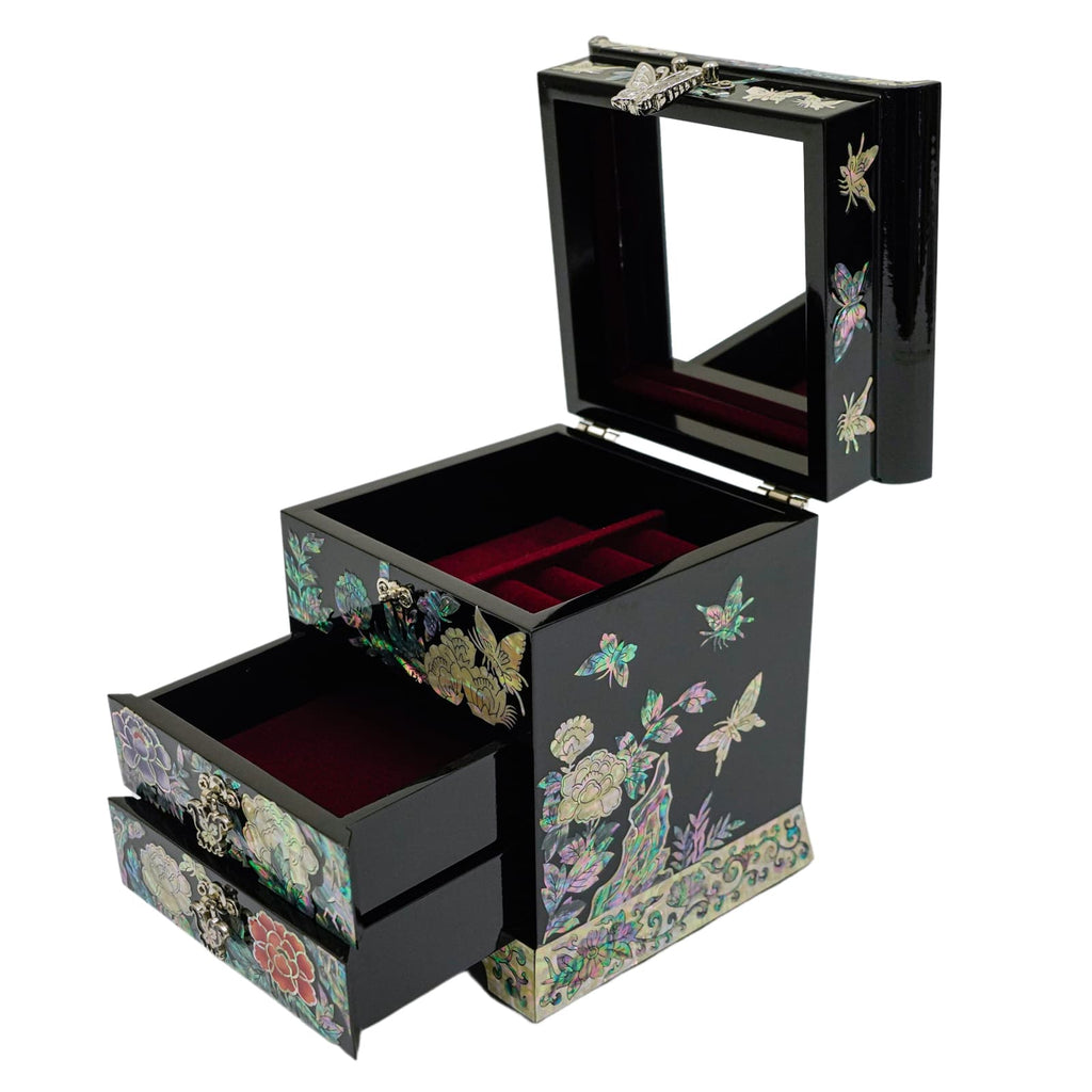 An open jewelry box with a mirror in the lid, velvet ring slots, and two large drawers with mother-of-pearl designs on a black background.